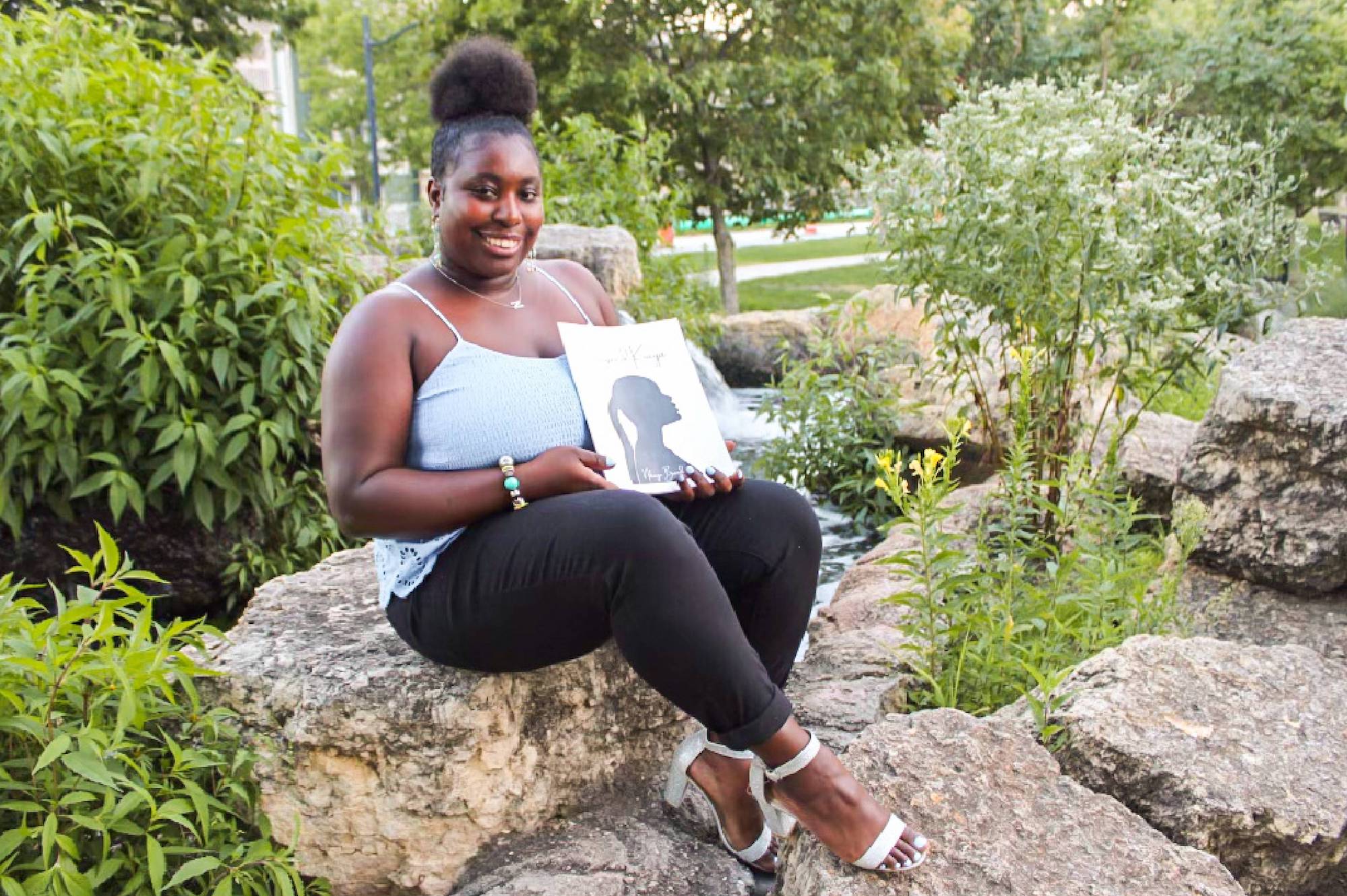 Nikiaya Brandon is a young Black woman. She wears black leggings, white sandal high heels, and a light blue spaghetti strap top. Her hair is in a poof on top of her head. She is sitting on a large rock outside at a beautiful park, holding her poetry book. The book is a large softcover. It has a white background with a profile silhouette of a Black woman, with her chin pointed upwards and a long brain hanging from a pony tail.