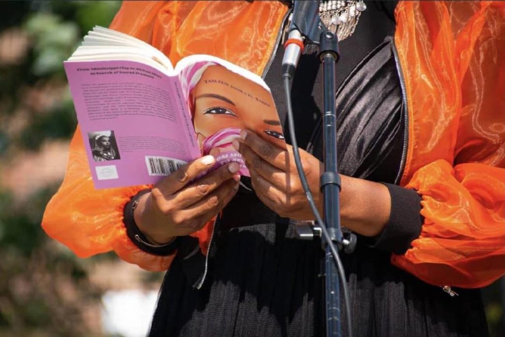 a close up of Black woman holding Tasleem Jamila's new poetry collection while standing in front of a microphone. You can only see her center and the book. She is wearing a black outfit with a bright orange jacket. The book has a pink cover with a close up shot of a woman's face. 