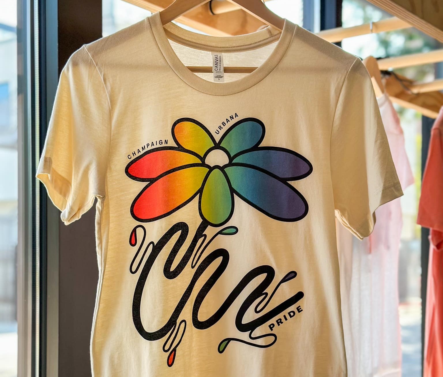 A cream colored t-shirt with a flower with rainbow petals hanging on a hanger.