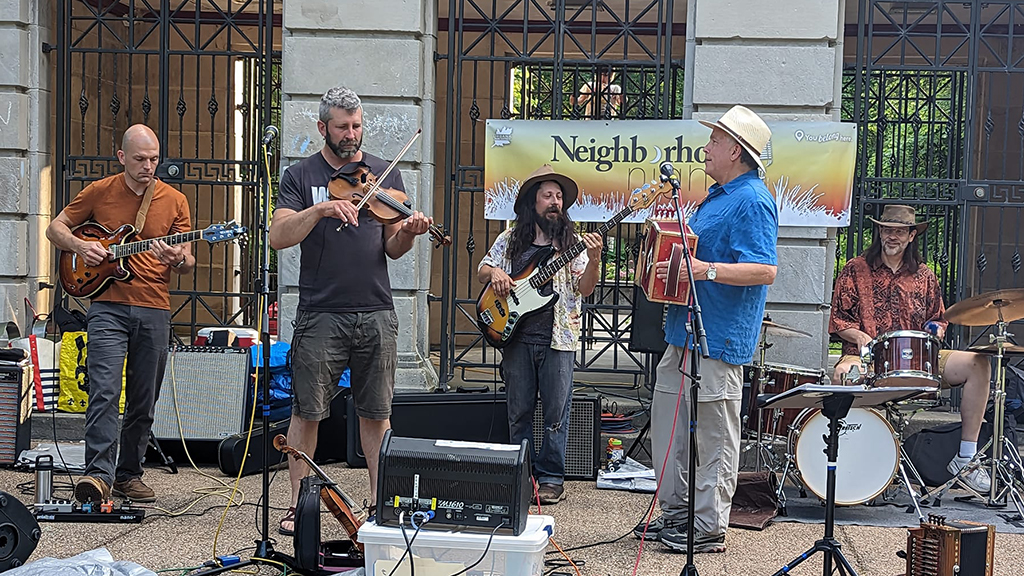 This is a photo of a band playing on a stage in an outdoor setting. The stage is set up in front of a large gate with a banner that reads “Neighborhood”. The band consists of five members, all of whom have their faces blurred. The band members are playing various instruments, including guitars, a violin, and drums. There are several amplifiers and other equipment on the stage. The background consists of trees and a building.