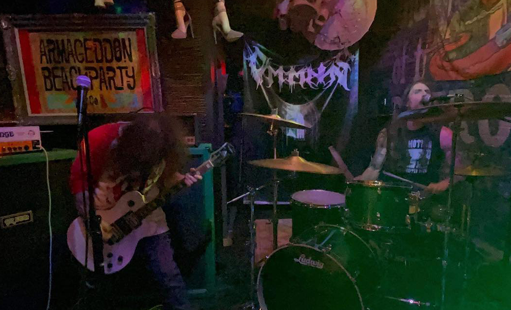 This is a photo of a band performing on a small stage in a dark bar. The stage is decorated with colorful graffiti and a banner that reads “Armageddon Beachparty”. The band consists of a guitarist and a drummer. The drummer is playing a green drum kit with a cymbal and a hi-hat. The guitarist is playing a white electric guitar and is standing in front of a black amplifier. There is also a microphone stand on the stage, but it is not in use.