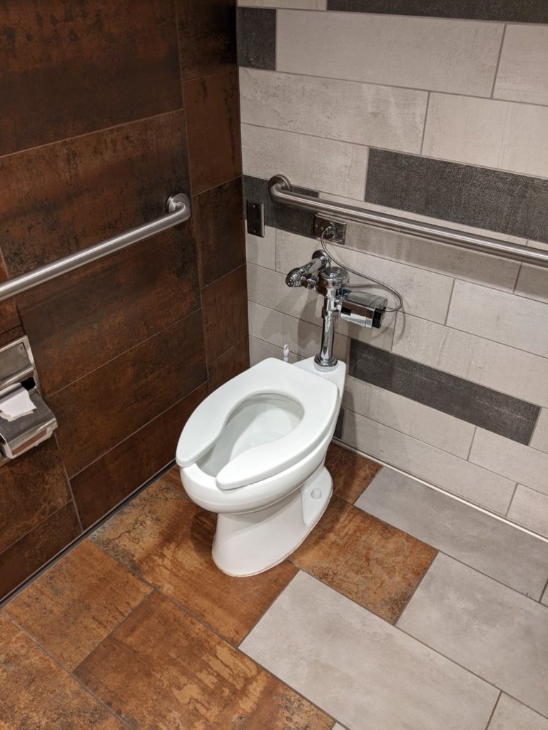 A dark gray and white tile wall next to a white toilet with a half wood and half tile floor.