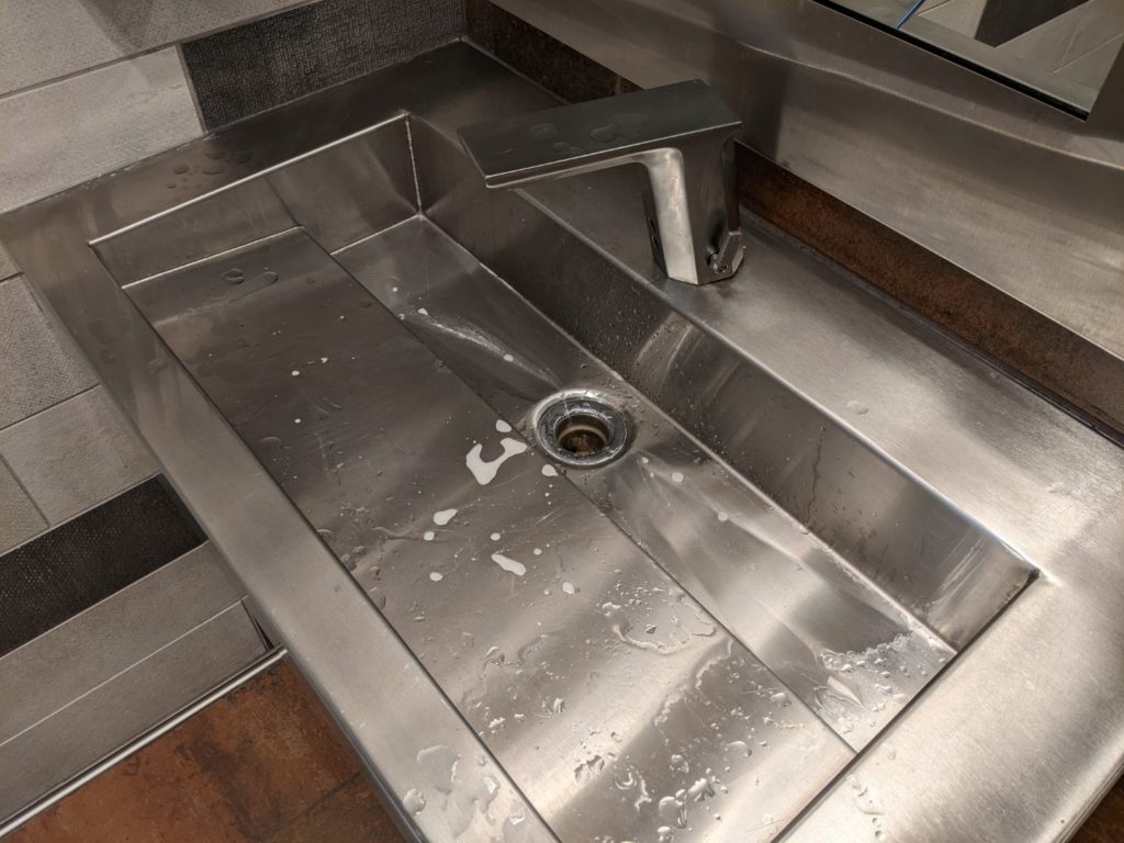 A big industrial metal sink with a silver flat edged faucet. A mirror is visible the top of the picture.