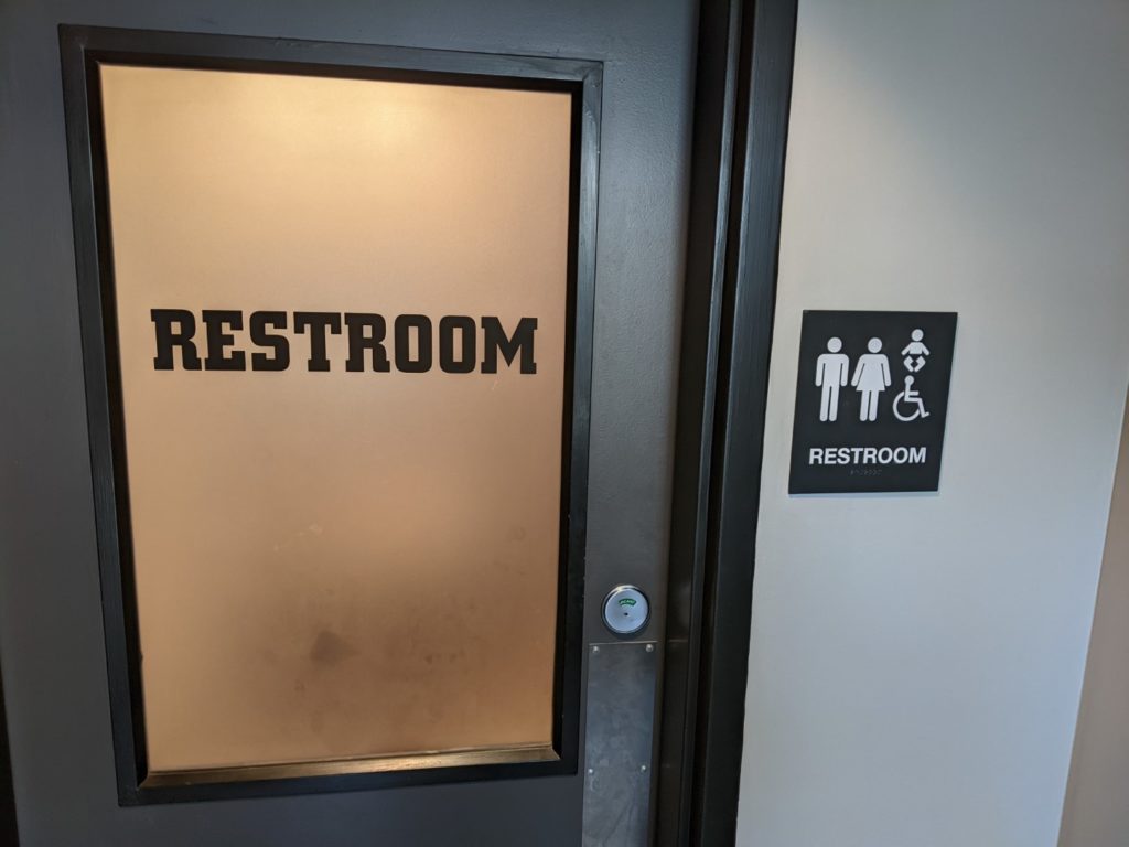 A silver door with a frosted window panel with the word restroom in big black letters. There is a sign on the wall to the left that shows it is an all gender restroom.