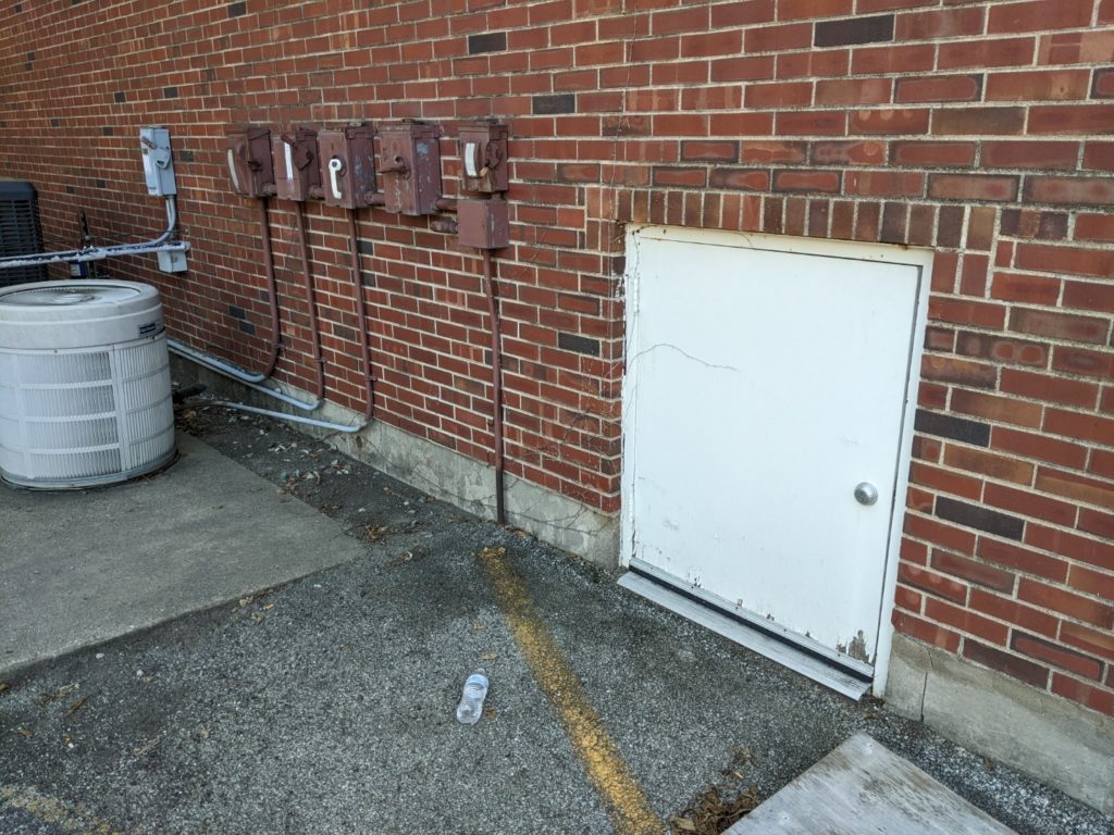 A brick wall with some meters attached to the wall and a very small white door. There is a parking lot visible in the lower left side of the picture.