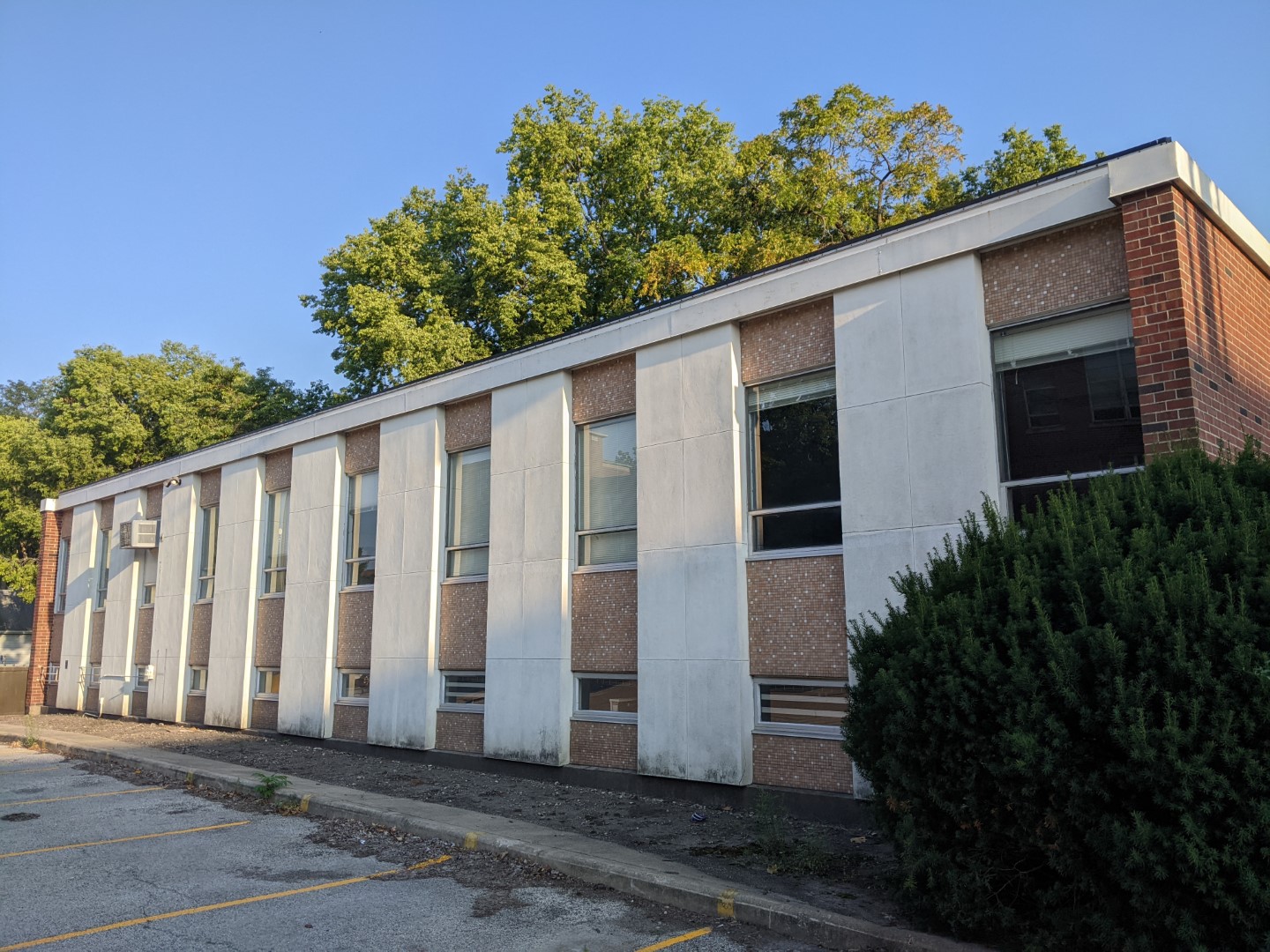 The outside of a brick and cement building. There are columns separating tile walls and windows. There is a parking lot visible in the lower left corner. It's a sunny day with no clouds.