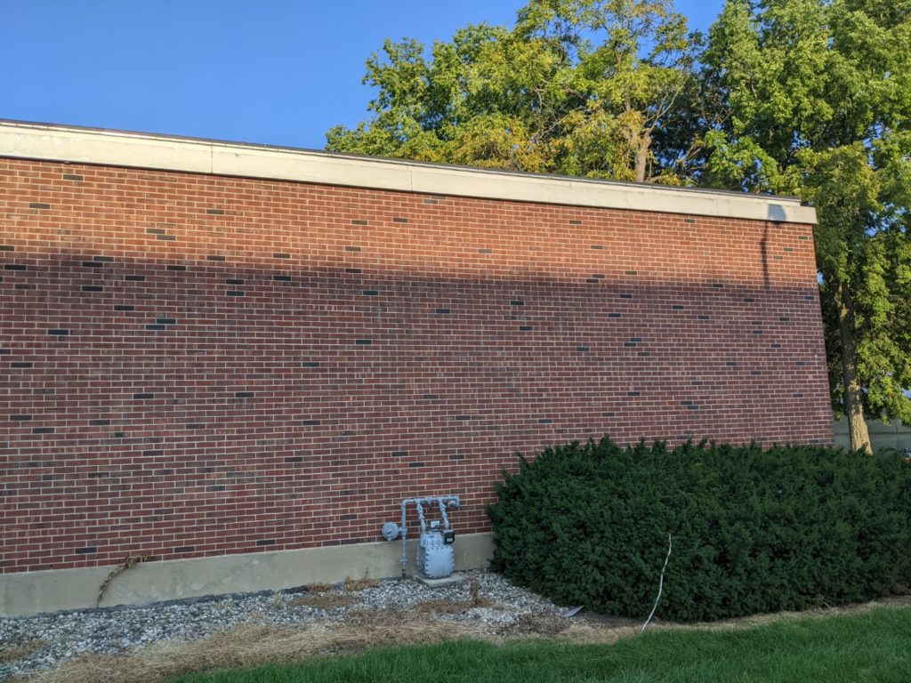 The backside of a brick building. Made up of a solid brick wall. There is a low green bush on the right side and trees and blue sky visible above the building. 