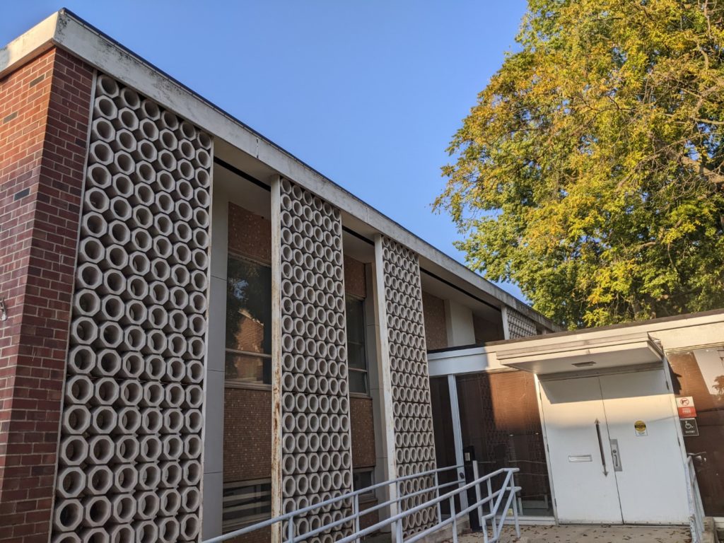 The outside of a brick and cement building. There are small cement circles, stacked up to create columns and a set of stairs leading down to a solid white door. It's a sunny day with no clouds. 