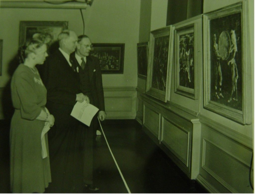 A black and white photo showing : Mrs. Stoddard, President George Stoddard (center), and Professor Frank Roos, Head of the Art Department, examining art hanging 