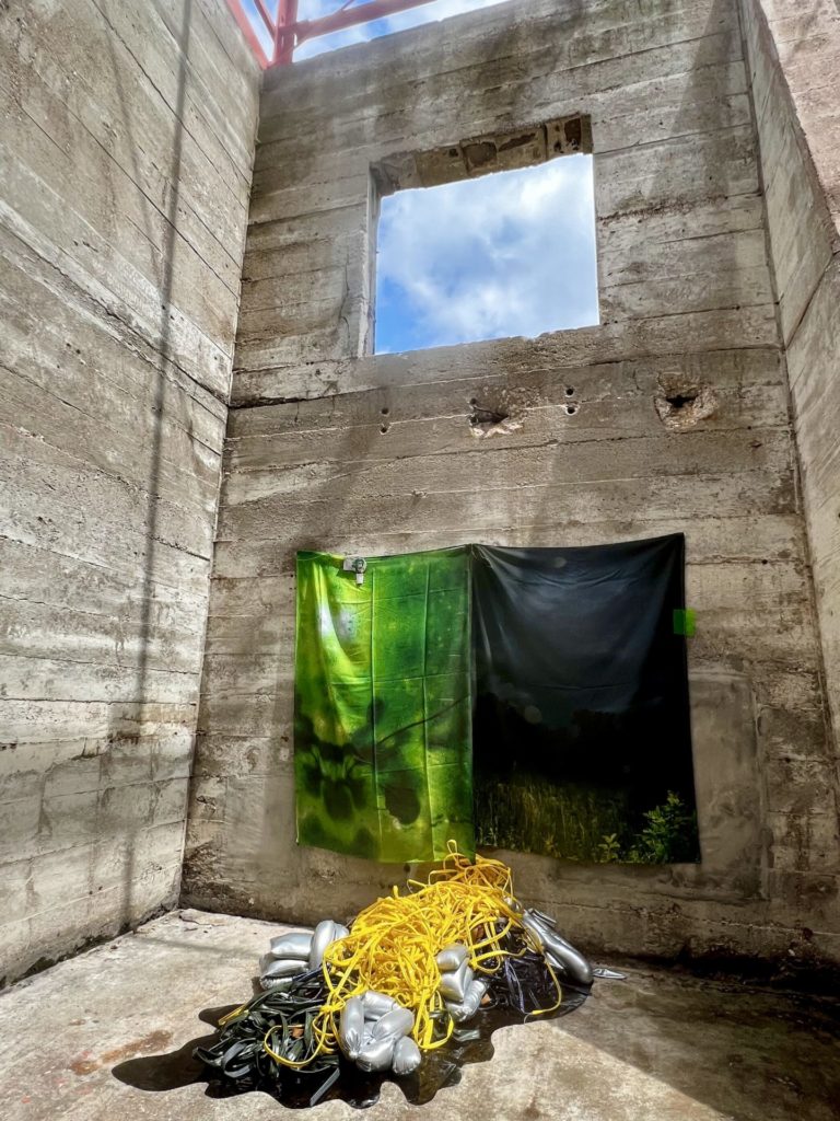 large rectangular fabric hang in an abandoned building. there is an open window above the fabrics. The fabrics are bright green on the left and dark navy black on the right. On the ground is a large bunch of cords and other supplies. 