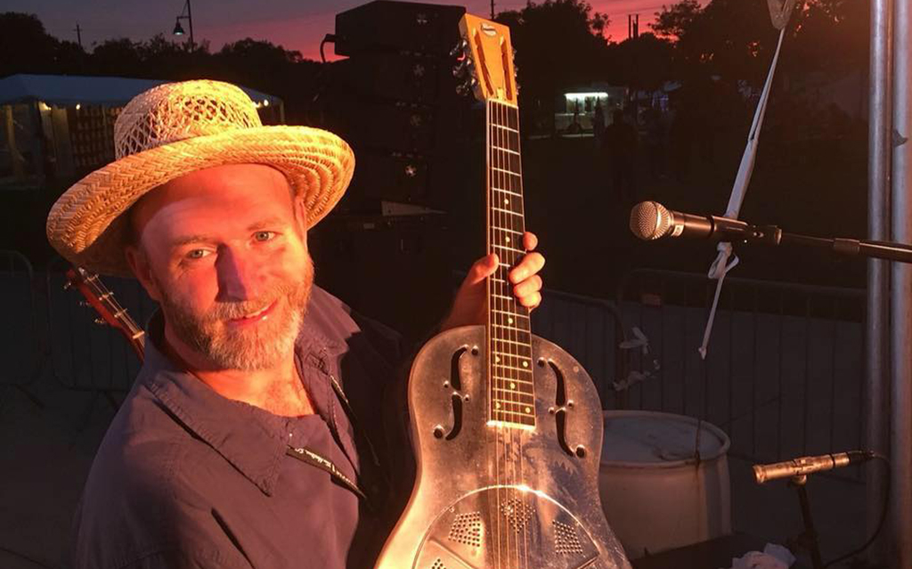 This is a photo of a person holding a guitar on a stage. The person is wearing a straw hat and a green shirt. The guitar is a silver resonator guitar with a yellow strap. The background is a sunset sky with a stage and some equipment visible. The photo is taken from the side of the stage.