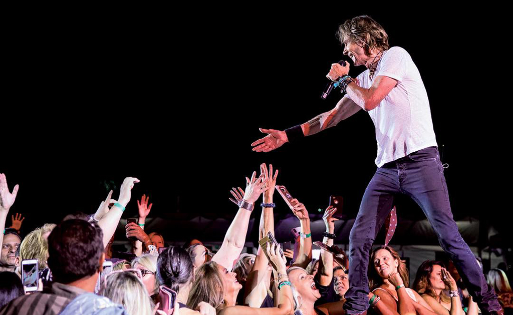 The image is a photo of a singer performing on stage in front of a crowd. The singer is wearing a white t-shirt and dark pants, holding a microphone and reaching out to the crowd, who are reaching back towards the singer. The background is dark, but you can see stage lights. 