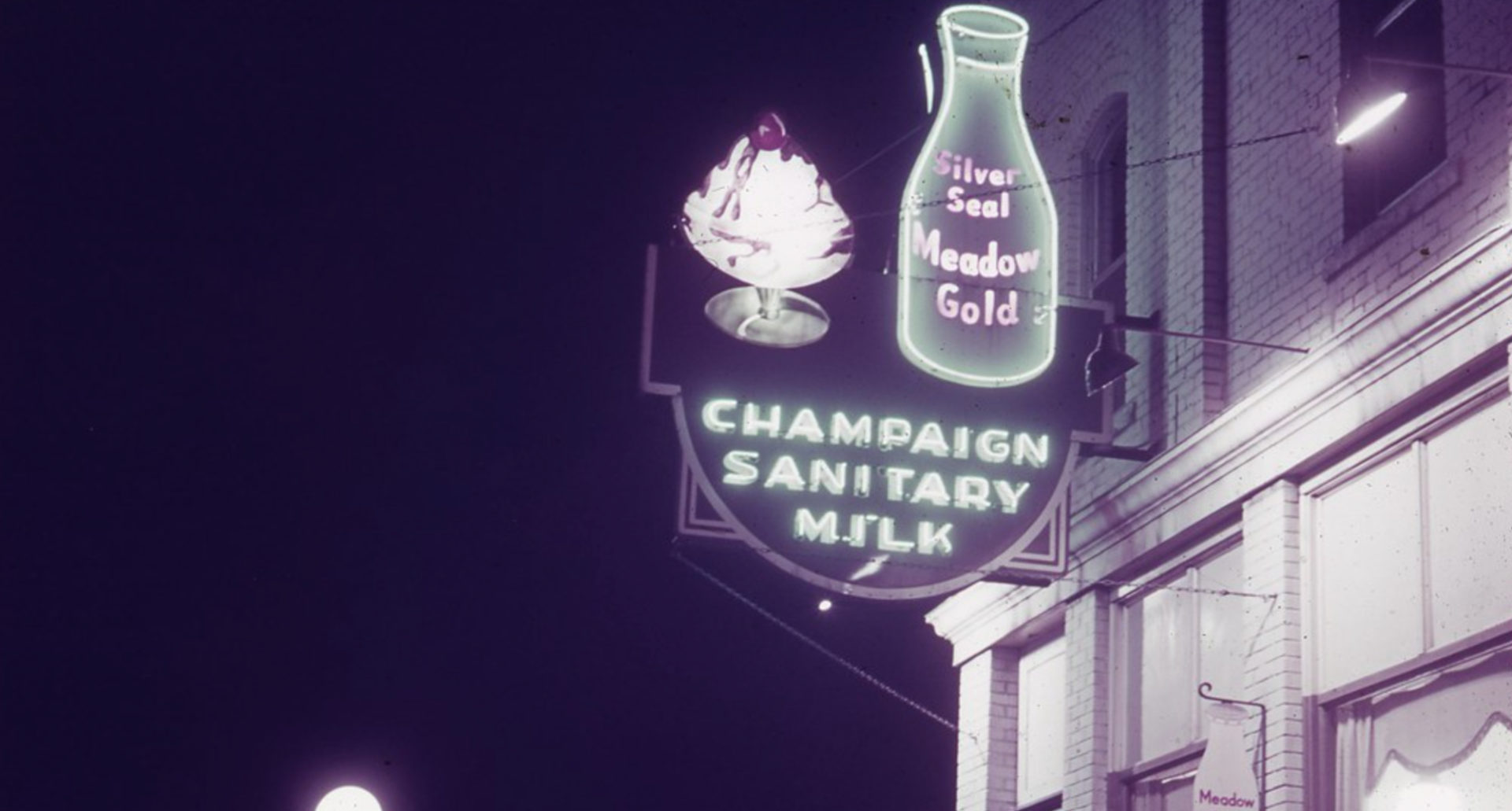 A neon sign in at 415 East University Avenue, Champaign in 1939 has an ice cream sundae, a decanter of milk, and below the words "champaign sanitary milk."