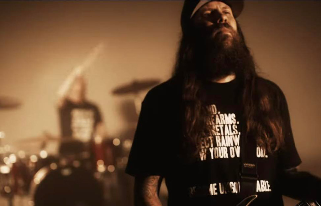 This is a photo of a person standing in front of a drum set in a dimly lit room. The person is wearing a black t-shirt with white text on it. The person has a long beard and long hair. The drum set is in the background and is black in color. The background consists of diffused lighting.