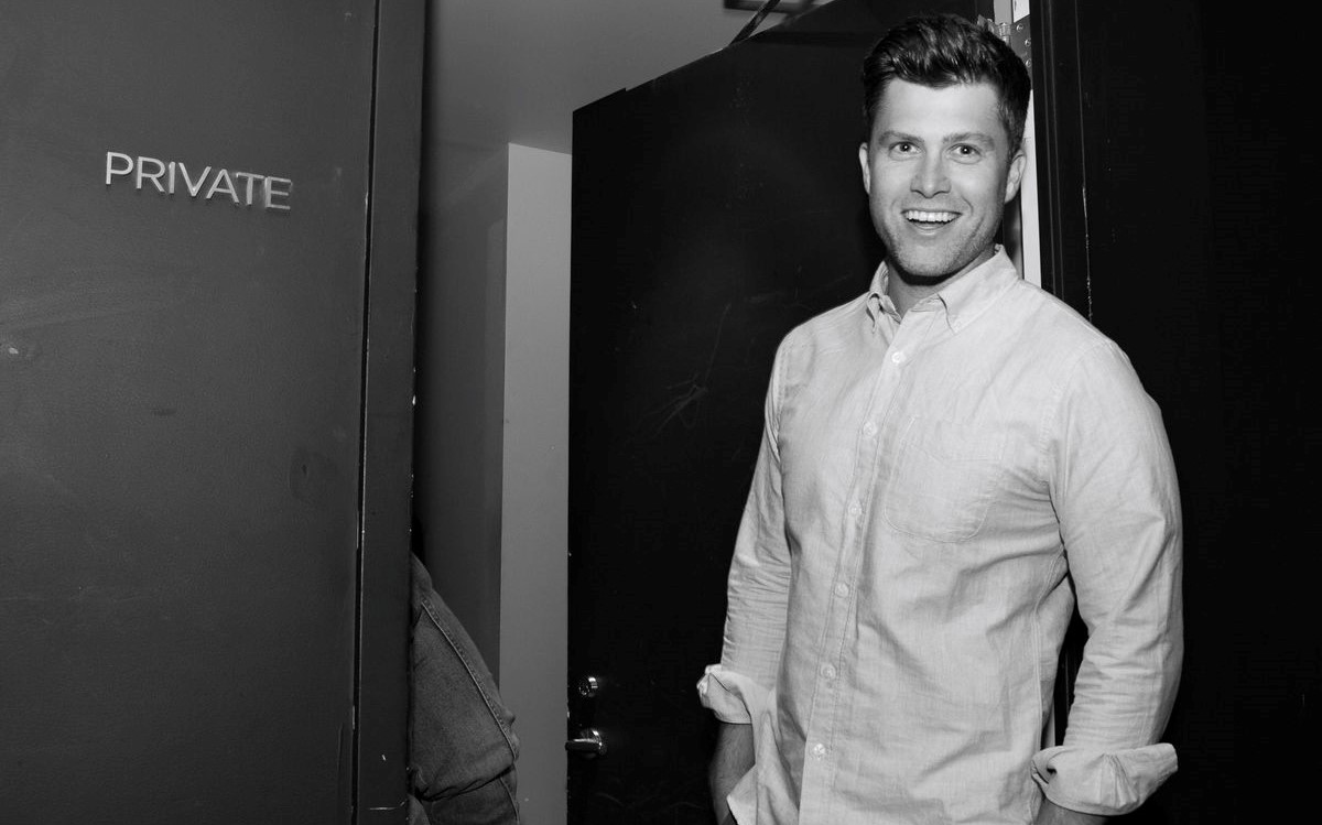 Black and white image of Colin Jost, a white man in a collared shirt standing in a doorway.