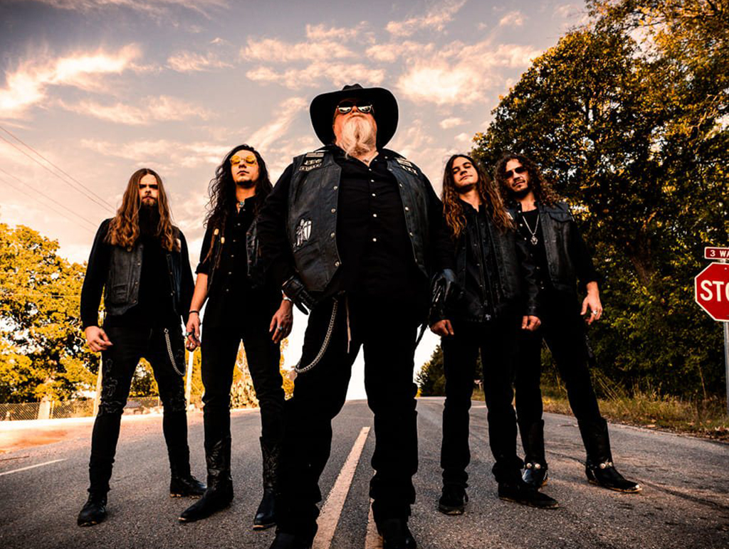 This is a photo-realistic image of a group of people standing on a road with a stop sign in the background. The group is dressed in black clothing and leather jackets. The person in the center is wearing a cowboy hat and has a long beard. The background consists of trees and a clear sky.