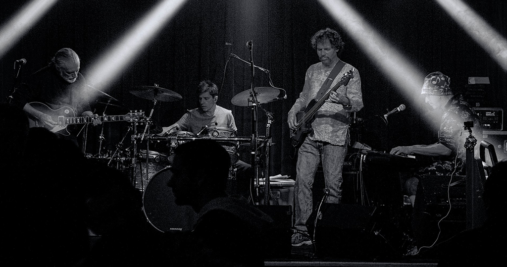 This is a black and white photo of a band performing on stage. The band consists of four members: a guitarist, a drummer, a bassist, and a keyboardist. The guitarist is on the left side of the stage, the drummer is in the center, the bassist is on the right side of the stage, and the keyboardist is on the far right side of the stage. The stage is lit with spotlights, creating a dramatic and high contrast lighting effect. The audience is visible in the foreground.