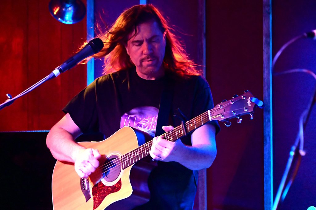 This is a photo of a person playing an acoustic guitar on a stage. The person is wearing a black t-shirt with a graphic on it. The guitar is a light wood color with a red pickguard. The background is a stage with red and blue lighting. There is a microphone in front of the person.
