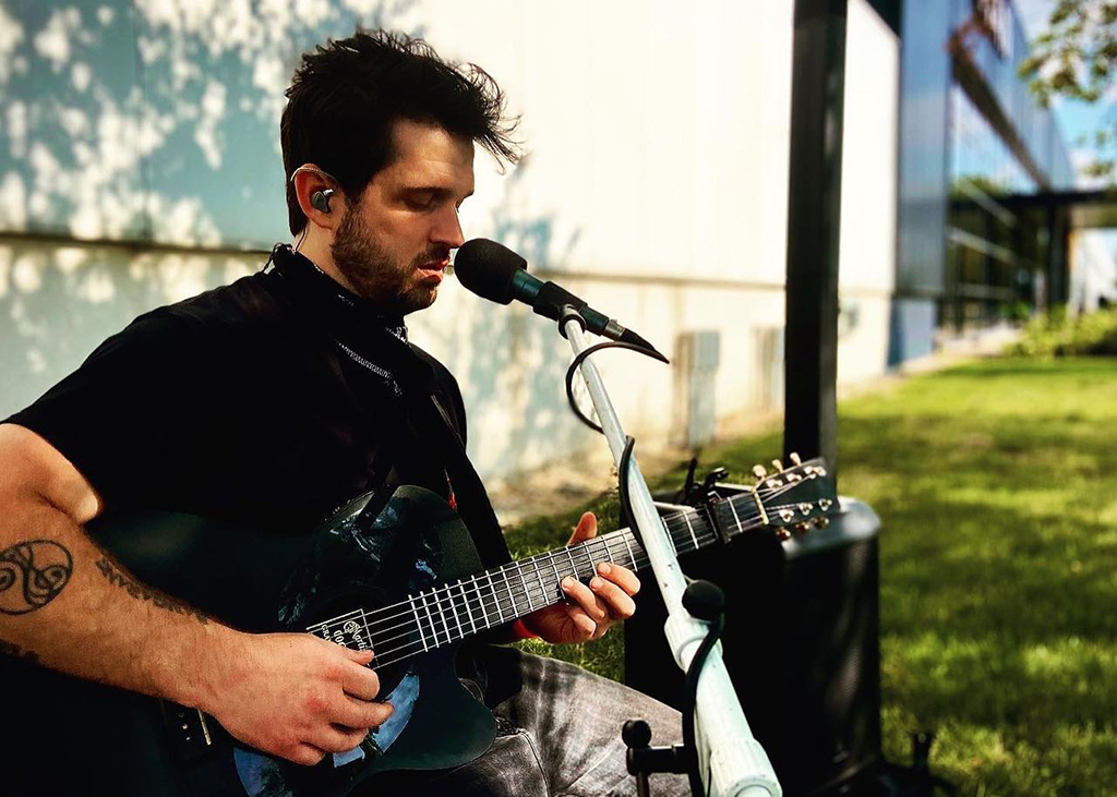 The image is a photo of a person playing an electric guitar in an outdoor setting. The person is wearing a black t-shirt and has a tattoo on their left arm. The guitar is black and white with a silver neck, and the person is also wearing earphones and singing into a microphone. The background consists of a grassy area with trees and a building in the distance.