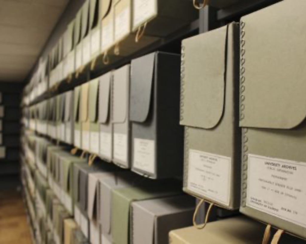 A view of the University of Illinois Archives. 4 levels of boxes in faded colors of blue and green sit on shelves in long rows along a wall. They all have identical white paper labels, listing the contents inside.