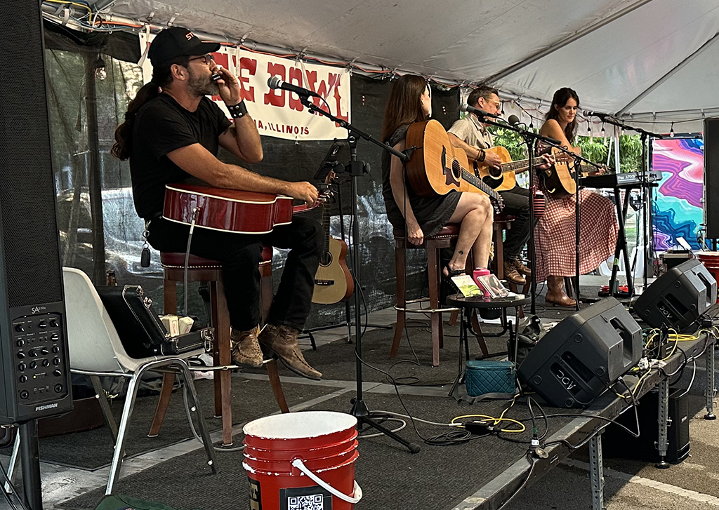 This is a photo of a band performing on a stage under a tent. The band members are sitting on chairs and playing guitars. The stage is decorated with a colorful banner and a large screen displaying abstract art. There are speakers and other musical equipment on the stage. The background consists of a white tent and trees.