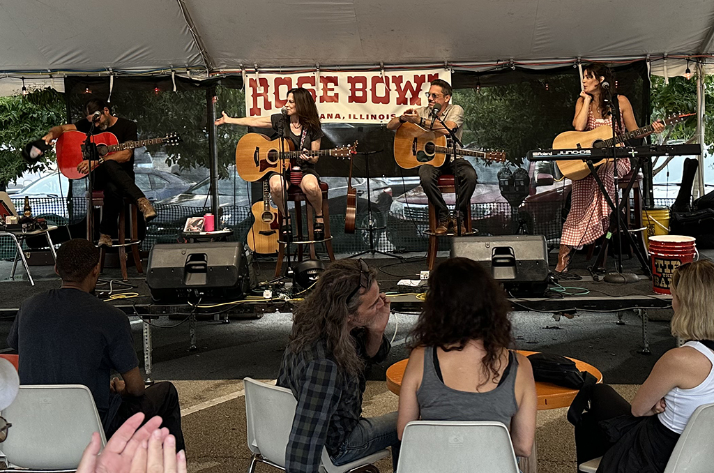 This is a photo of a band performing on a stage at an outdoor event. The stage is set up under a white tent with a banner that reads “ROSE BOWL” in red letters. The band consists of four members, two playing guitars, one playing a keyboard, and one playing a drum. There are people sitting in white folding chairs in front of the stage, watching the performance. The background consists of trees and a parking lot.