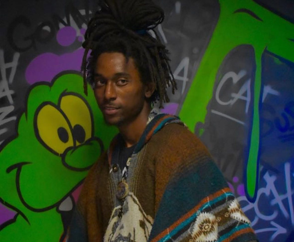 This is a photo of a person standing in front of a colorful graffiti wall. The person is wearing a multicolored sweater with an Aztec-like pattern and has dreadlocks. The graffiti wall has a green cartoon character and various tags and signatures in different colors. The background is dark and the lighting is low.