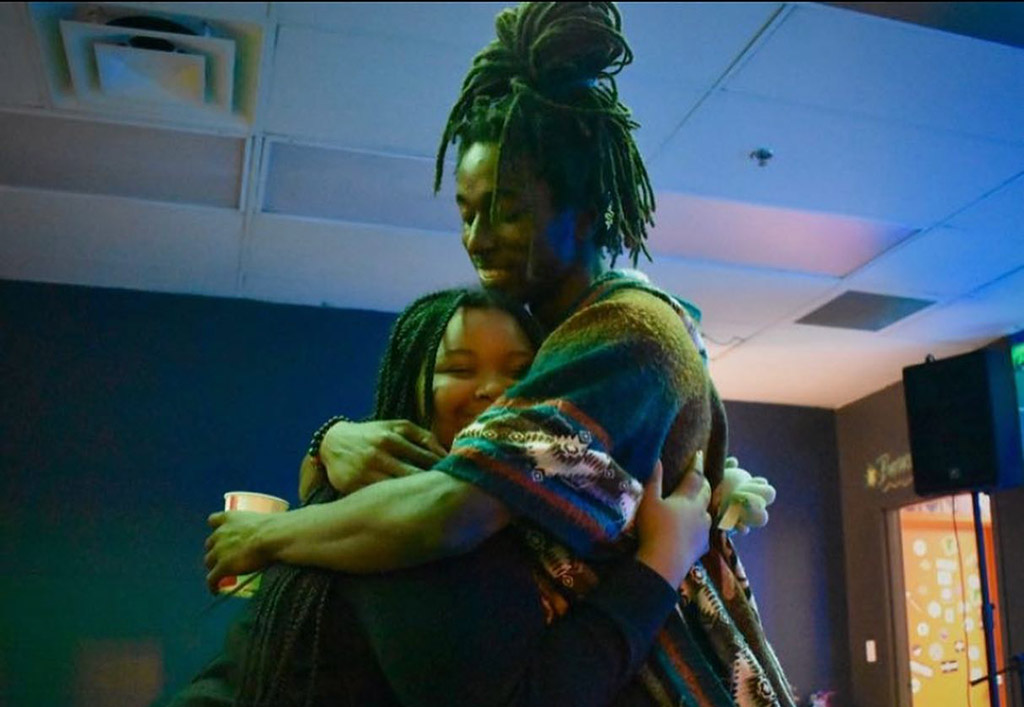 This is a photo of two people hugging in a room with a blue-green wall and a ceiling vent. The person on the right has long dreadlocks and is wearing a colorful scarf. The person on the right is holding a red cup in their hand. There is a speaker in the background on the right side of the image.