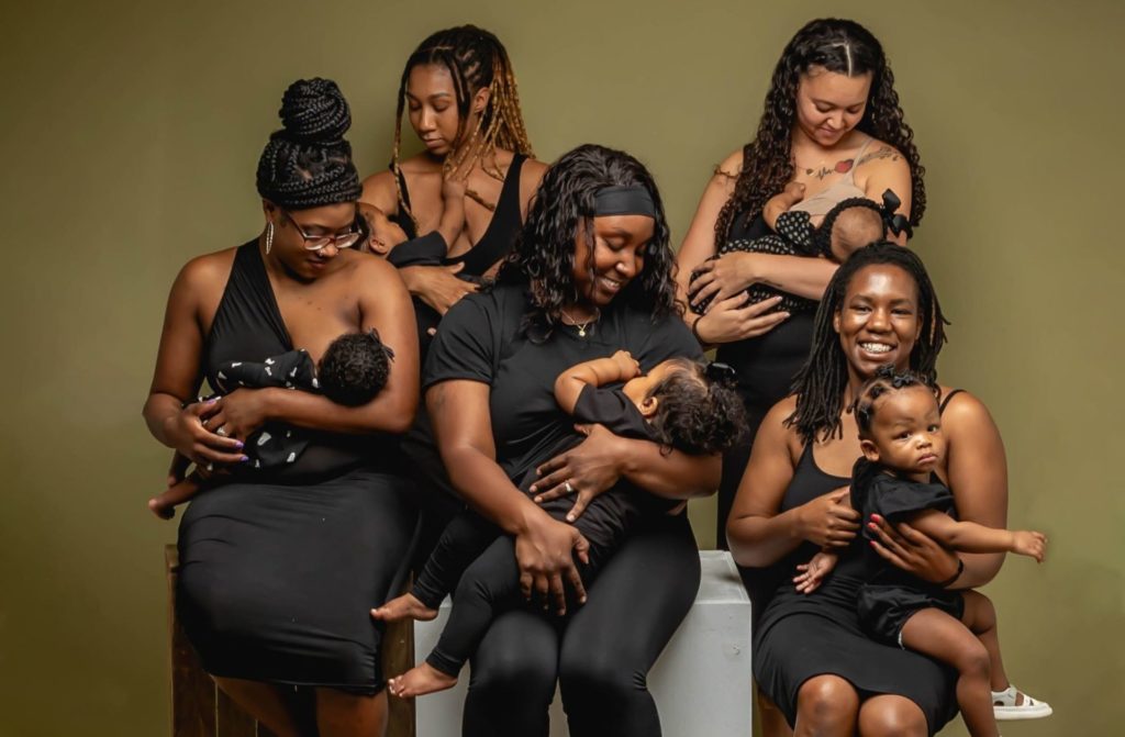 A group of Black and Brown woman are dressed in black and and gathered for a photo while breastfeeding their children.