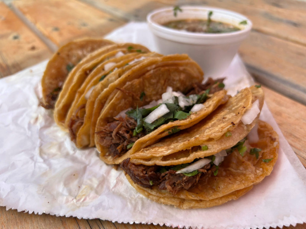 Five birria tacos with cilantro and white onion are on a white parchment paper on top of tin foil. There is a white styrofoam cup of consume behind the tacos. Photo by Alyssa Buckley.