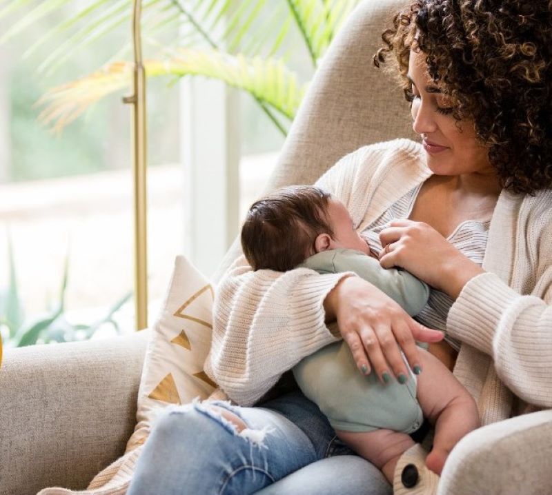A woman with brown curly hair is seated in an armchair, breastfeeding an infant.