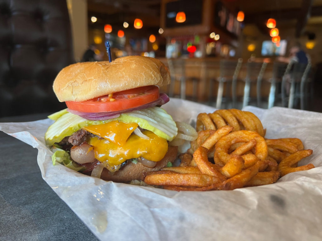 A Western burger with American cheese, bacon, tomato, red onion, pickles, and two beef patties on a bun with a blue-tipped toothpick. Photo by Alyssa Buckley.
