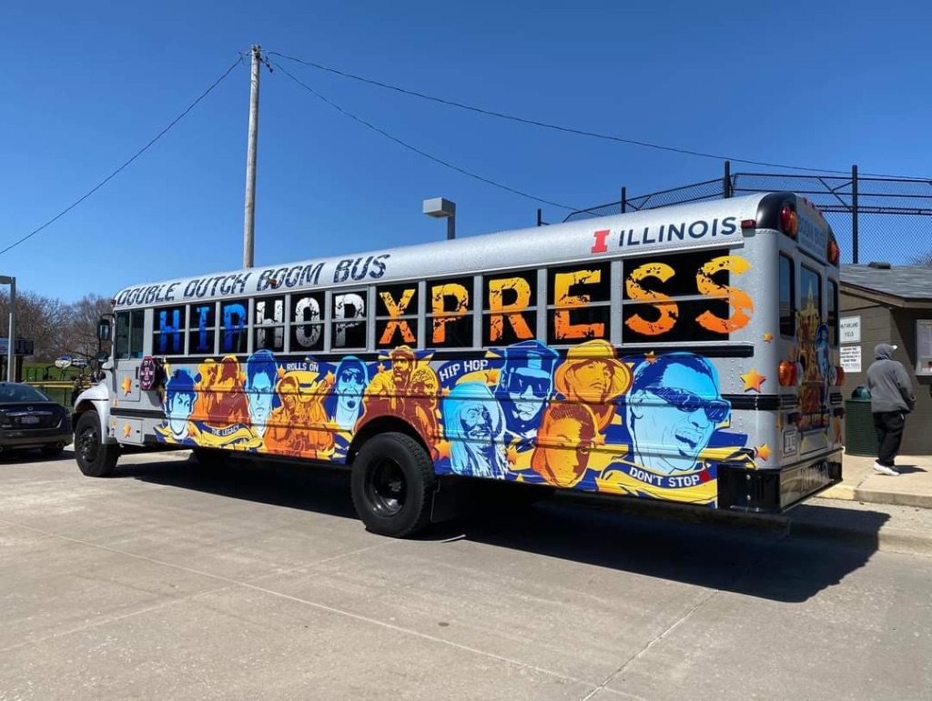 A large gray school bus is painted with a colorful blue and orange mural. It is sitting In the parking lot