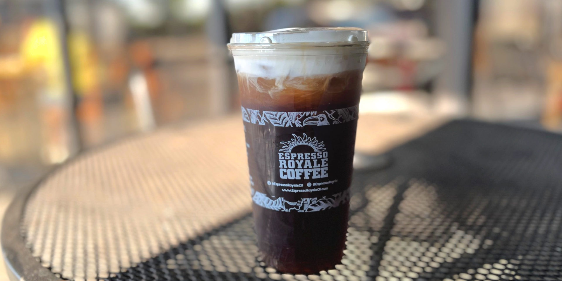 Espresso Royale at Stadium Plaza's Americano with cold foam is in a to-go cup with the coffee shop's logo on the outside on a black patio table outside the coffee shop in Champaign. Photo by Alyssa Buckley.