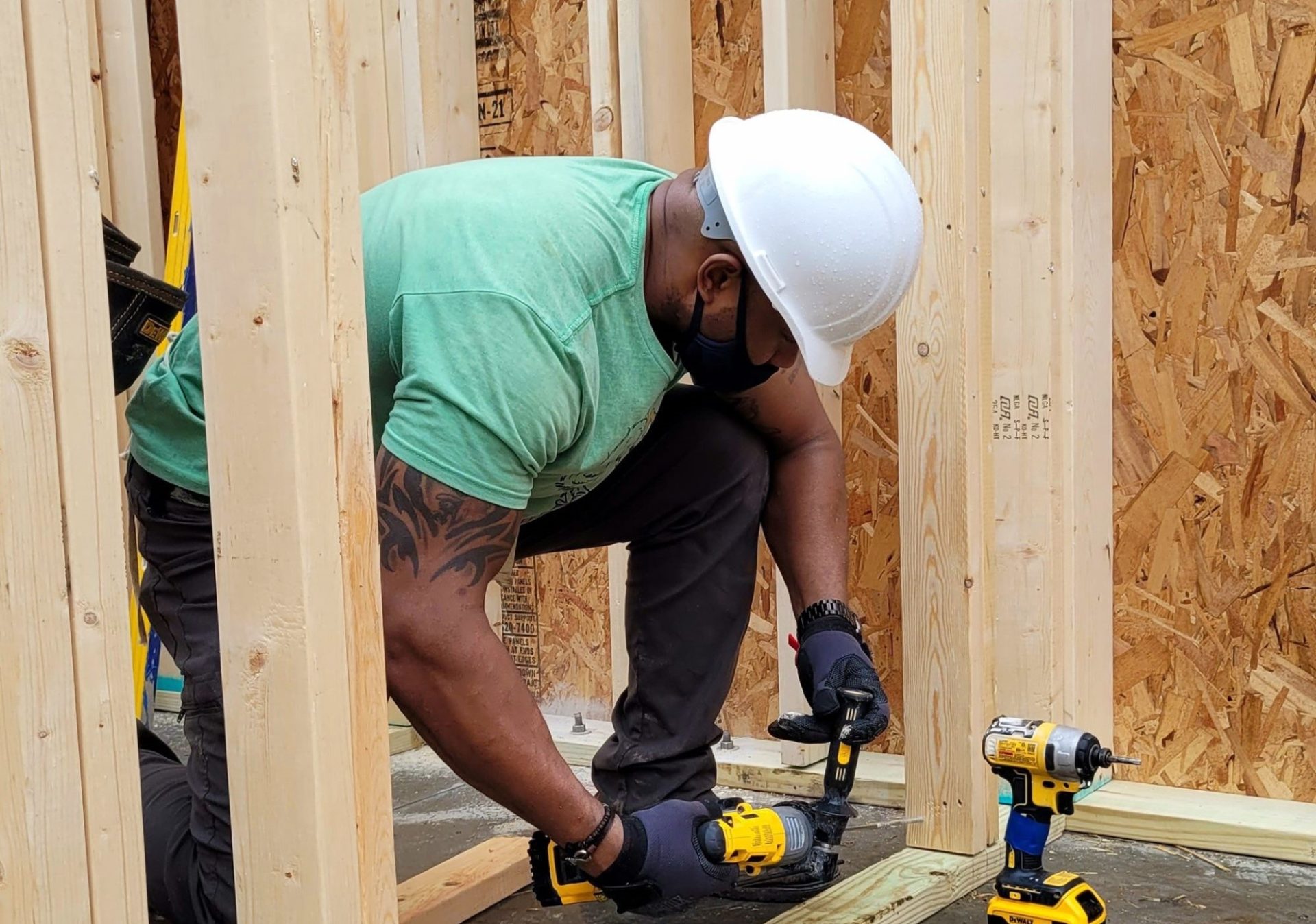 A Black man in a mint green shirt and white construction helmet is crouched down, hammering a nail.