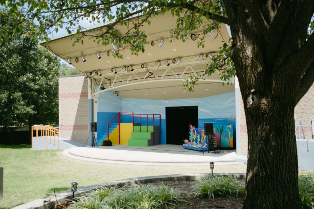 An outdoor stage with colorful blue backdrop, and a large grassy area in front. There is a large overhang that is lined with stage lights. 