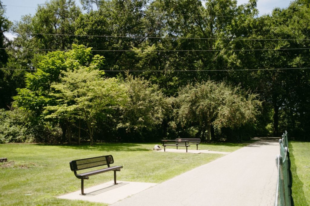 A paved trail stretches into a wooded area, with grass on either side. There are metal benches on one side, and a green fence on the other.
