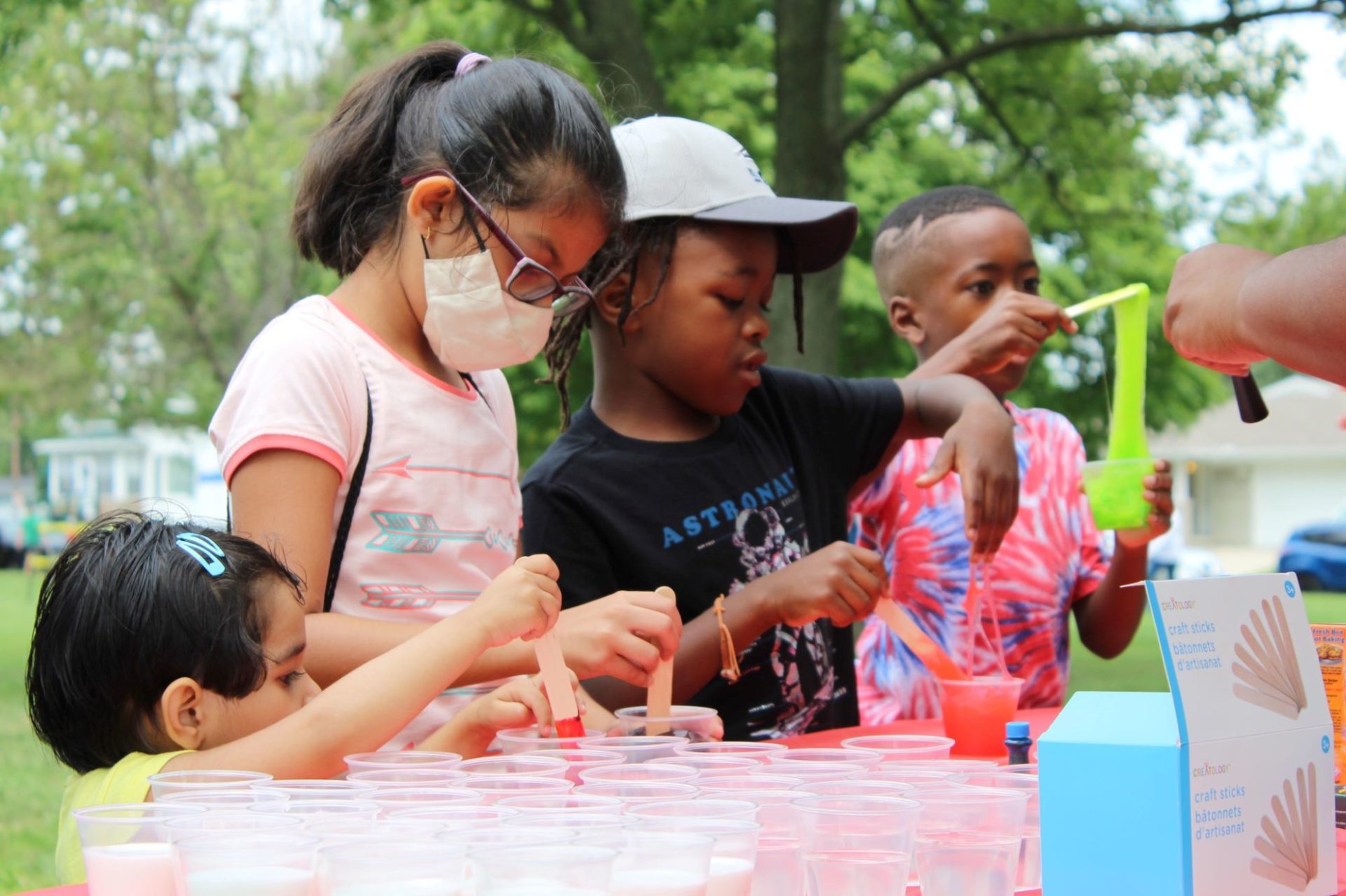 Four kids are lined up along a table with multiple empty clear containers. They are each mixing colored slime in a container.
