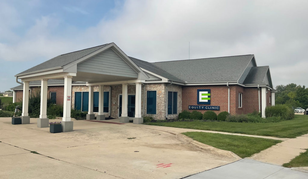 Equity Clinic is an abortion provider in Champaign. The brick building has stone detailing and covered entrance extending to the driveway. The sign is blue, white, and bright green, and says Equity Clinic.