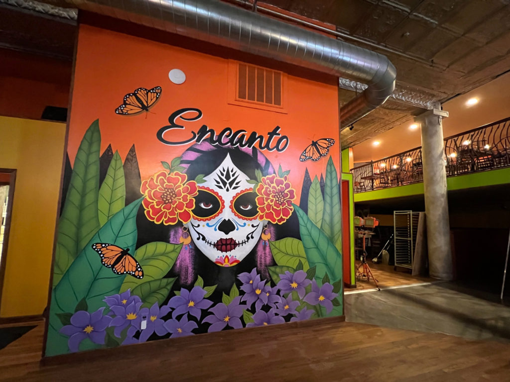 Inside Encanto Restaurant and Bar, there is a mural with monarch butterflies and a feminine face with Day of the Dead makeup poking out of long leaves and purple flowers. Photo by Alyssa Buckley.