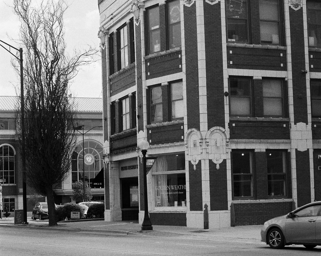 A black and white picture of the outside of the golden weather goods store. The building is older and made with a bit of bricks and cement. There are three floors all with windows overlooking the street out front.