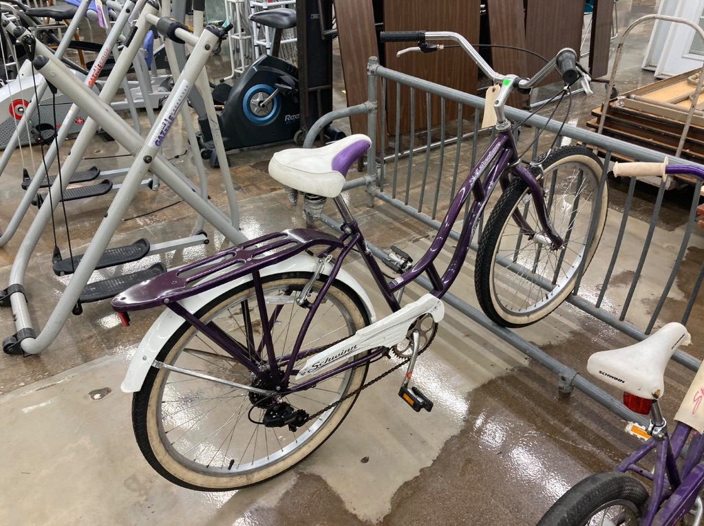 A purple and white bike is parked in a bike rack inside a store. It has a white seat and is surrounded by various equipment for sale. 