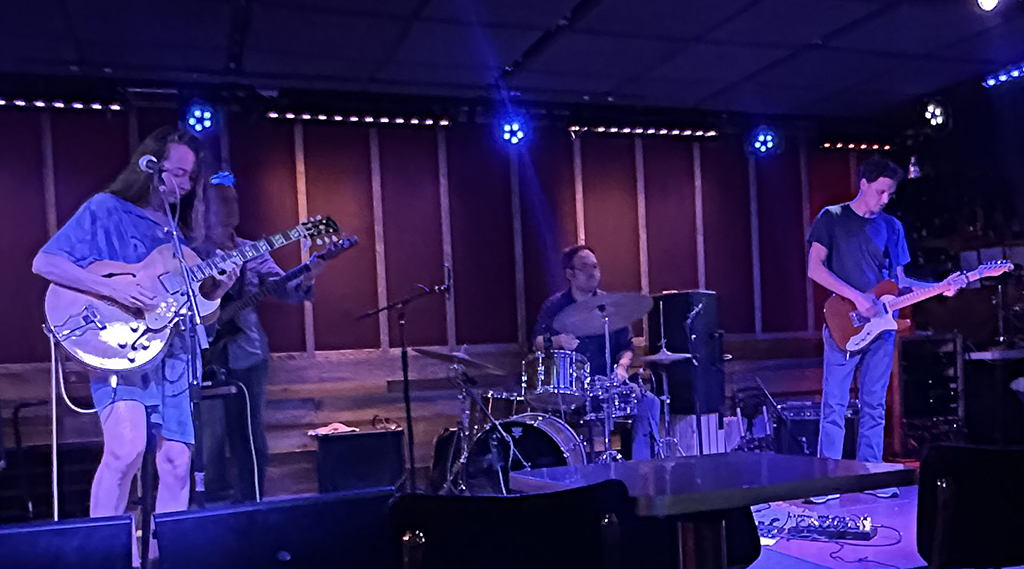 This is a photo of a band performing on a stage in a dimly lit venue. The band consists of a guitarist, a drummer, and a bassist. The guitarist is playing a white electric guitar and is wearing a blue Hawaiian shirt. The drummer is playing a drum kit with a cymbal and a hi-hat. The bassist is playing a blue electric bass guitar and is wearing a blue t-shirt. The stage is lit with blue lights and has a black curtain in the background. The venue has a wooden floor and a seating area with tables and chairs.