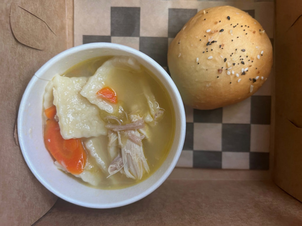 Inside a brown box, there is a cup of chicken noodle soup worth the drive to Mahomet for Lucky Moon Pies and More with a dinner roll dusted with sesame seeds. Photo by Alyssa Buckley.