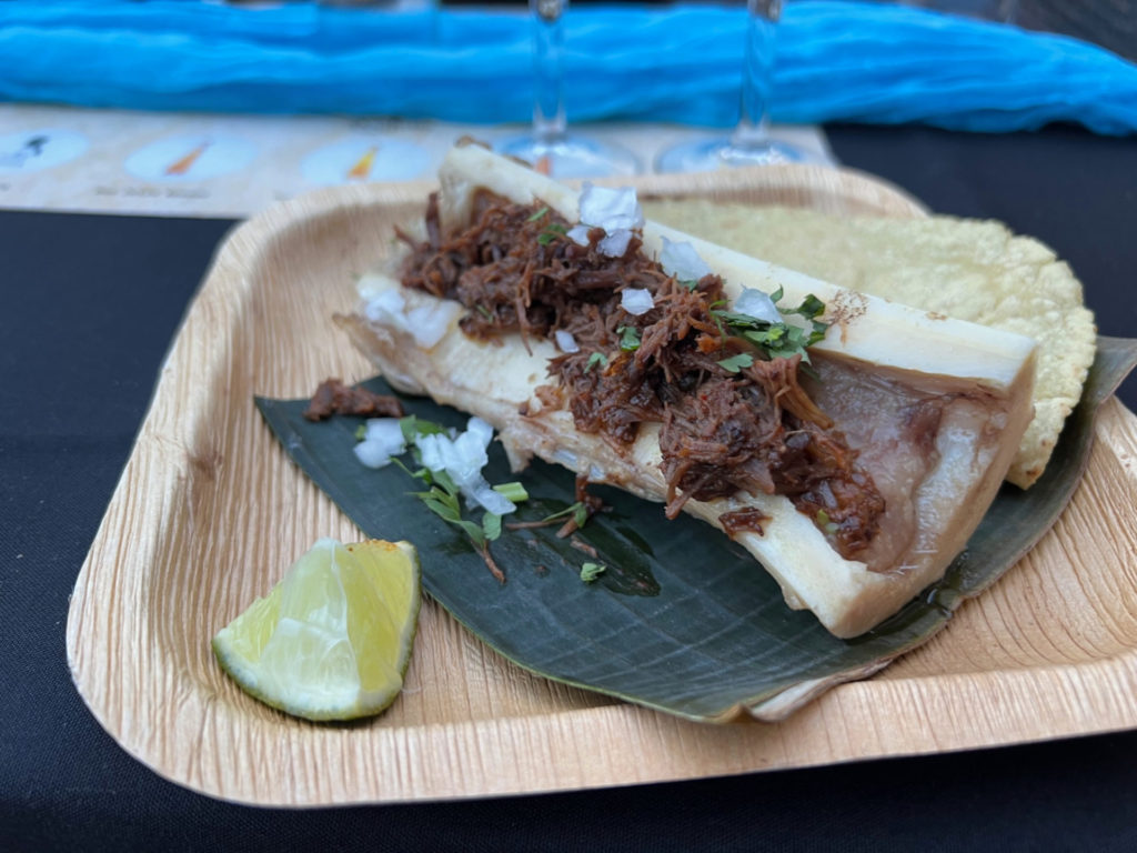 On a brown plate, there is a horizontally sliced bone with bone marrow topped with birra beef, cilantro, and onions. There is a wedge of lime and two tortillas. Photo by Alyssa Buckley.
