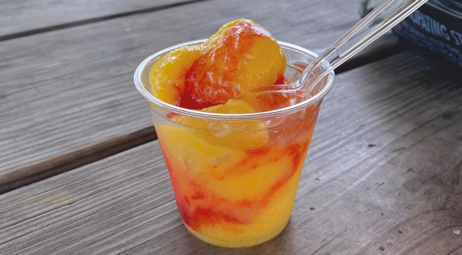 A cup of frozen mango dessert with a plastic spoon, sitting on a wooden picnic table.