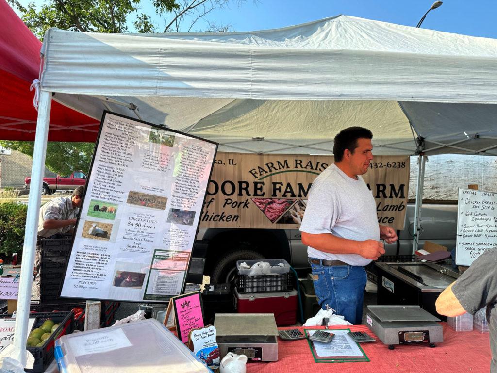 A man, Wes Moore, stands behind a table in front of a banner that says Moore Family Farm. He is wearing a light gray shirt and blue jeans. To his right, there is a freezer containing several cuts of meat. In front of him are two scales, and a sign that lists all of the meats that Moore Family Farm sells. Photo by Jake Williams.