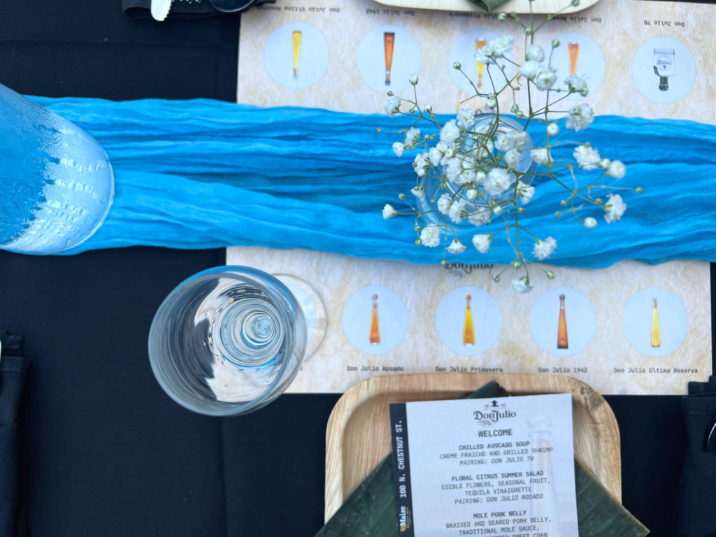 An overhead photo of the table at Maize's tequila dinner shows the Don Julio tequila tasting mat with the first glass of Don Julio 70 poured. There is a blue runner with a vase of baby's breath flowers. Photo by Alyssa Buckley.