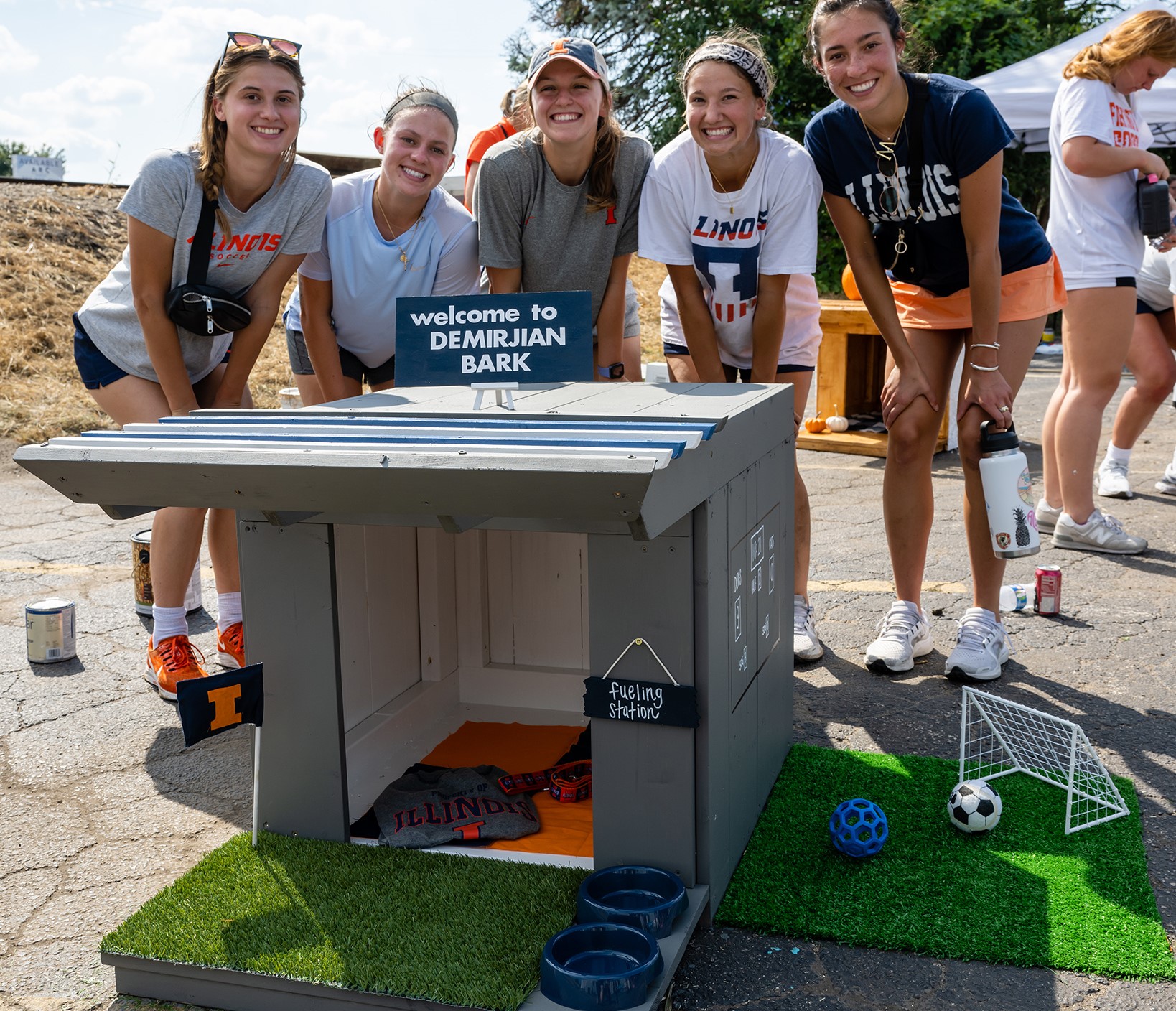 Five young white womean are gathered around a dog house that is painted gray and sitting on a green turf platform. There is a mini white soccer goal on the turf, and two small balls.