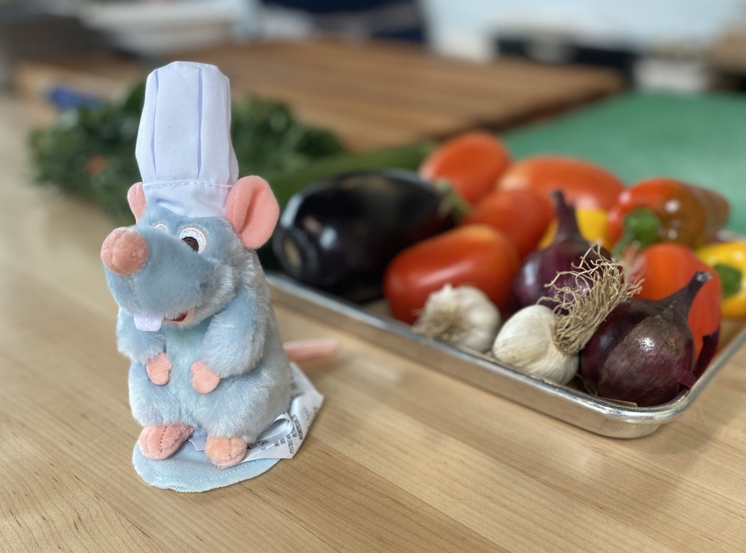 A metal tray of colorful vegetables on a wooden counter. A small stuffed mouse in a chef's hat is in the foreground.